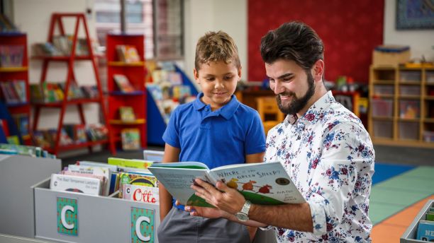 A male teacher reads a book to a young student. 