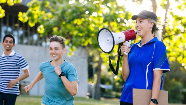 A female sport teacher speaks into megaphone while two students run. 